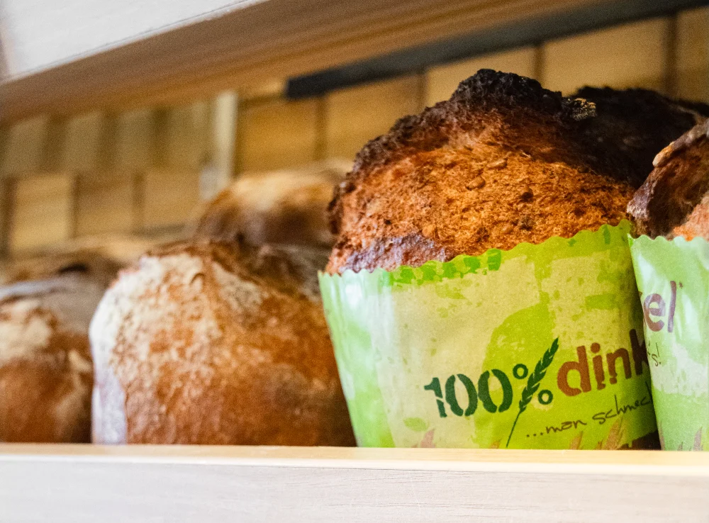 Nahaufnahme von frisch gebackenem Dinkelbrot auf einem Holzregal, wobei eines in grünes Papier eingewickelt ist, auf dem "100%" und andere teilweise sichtbare Texte aufgedruckt sind.