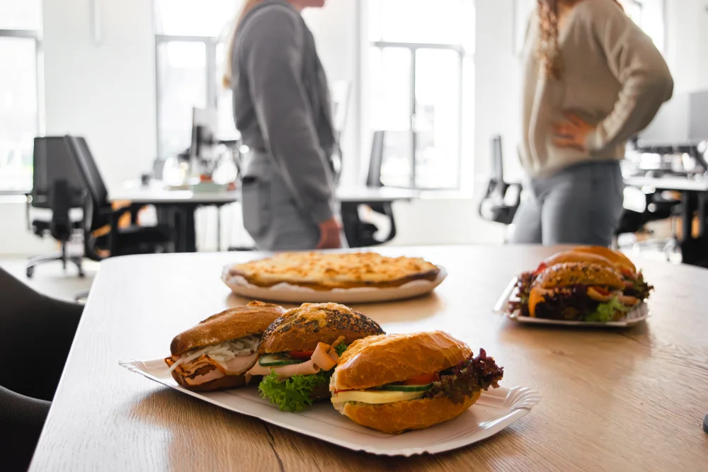 Auf einem Tisch in einem Büro liegen Sandwiches und ein Hefekuchen. Im unscharfen Hintergrund, in der Nähe heller Fenster und leerer Schreibtische, stehen zwei Personen und unterhalten sich.